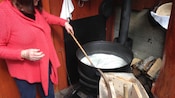 A woman with wooden spoon stands above a large pot stirring milky contents