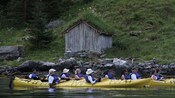 A group of people in kayaks floats past a wooden shack with grass on the roof.