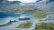 A building sits in front of a lake and gravelly hillside peppered with sparse vegetation