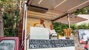 A food truck with 2 servers and a sign that reads Welcome Benvenuto Mickey Mouse Topolino