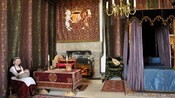 A woman dressed in historical clothing sits in a room of tapestries, including a coat of arms