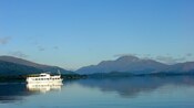 A boat with 2 decks cruising across a large body of water with hills in the distance