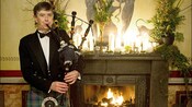 A man in a kilt plays bagpipes by a lit fireplace
