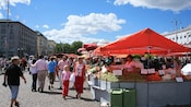 Crowds walk on cobble stone street by a variety of produce stands, in Helsinki