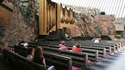 People sitting in pews at the Temppeliaukio Rock Church, featuring exposed brick walls and organ pipes
