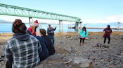Un hombre habla con un grupo sentado sobre rocas grandes mientras otros se paran y buscan pistas en una playa rocosa