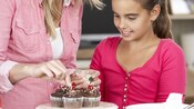 A girl watches as a woman sprinkles toppings on chocolate frosted cupcakes with cherries on top