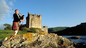 A bagpiper wears a tuxedo with a kilt, playing in front of an old castle on a barnacled hillside
