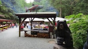 Un sitio para acampar con una parrilla al aire libre cubierta, mesas de pícnic y un letrero que dice “Salmon Bake” (Salmón al horno)