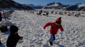 Rows of igloo shaped dog houses accompanied by sled dogs sit on a snow field as children play between them