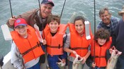 Four kids holding fishing rods and fish on the back of a boat smile into a the camera