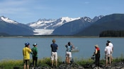 People standing by bicycles look at a nature vista featuring a large body of water, hills and snowcapped mountains