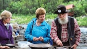 A guide dressed as an old prospector shows 2 women how to find gold in their mining pans