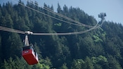 A cable car is suspended above the tree line from one of two cables hanging from a tower in the distance