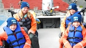 Um adulto e 3 crianças vestidos com coletes salva-vidas e equipamentos para pesca sentados na parte de trás de um barco