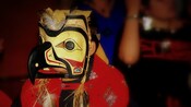 A child wears a large totem mask with a birds face