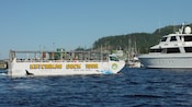 A Ketchikan Duck Tour land and sea vehicle is on the water near a boat and a dock