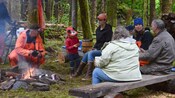 A group of 7 in wet weather gear sitting around a campfire
