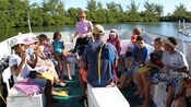 A group of children sit in the back of a boat in a half circle around an instructor carrying snuba gear
