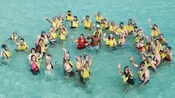 A large group of Guests wearing snorkeling gear wave as they wade in waist deep water
