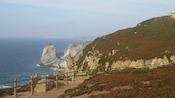 A wood fence runs along a dirt path on a cliff, overlooking the coastline