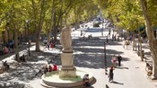 A statue and a fountain near people, a street, trees and storefronts