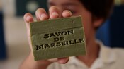 A person holds up a block of soap with words that read Savon De Marseille