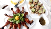 A plate of gourmet barbecue chicken drumsticks, fish fillets, rice and plantains and various sauces set on a table with a linen table cloth