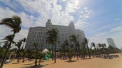 Un hotel enorme cerca de una playa con personas, árboles, sillas y un gazebo