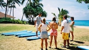On grass, 4 novice surfers watch as an instructor shows them the proper way to stand on a board