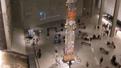 A steel beam from the twin towers stands vertically in a room surrounded by benches and people milling about