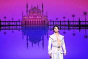 A stage scene of Aladdin depicts Aladdin standing in front of the Sultan’s Palace