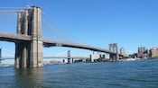A suspension bridge with two towers topped by American flags stretches across a river between cities
