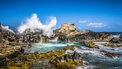 3 guests enjoy the Natural Pool while a wave crashes behind them on the protective boulders that surround the calm waters