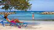 Una mujer reclinada en una silla de playa debajo de la sombra de un árbol cerca de un hombre sonriente que tiene el agua hasta la rodilla