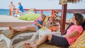 A family lies on lounge chairs and stand next to a wooden railing near the ocean