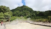 Hills and lush vegetation surround a wooden walkway over a series of steaming fumaroles