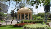 A domed gazebo supported by pillars in the middle of a plaza near trees and grassy lawns