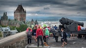 A group of people stand near a cannon on ramparts near a city
