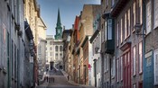 Tourists walk up a steep incline along a narrow street towards a steeple