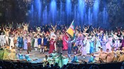 A group of performers dressed in 19th century attire on a stage with fireworks and fountains of water