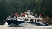 A 2 deck whale watching boat carrying passengers across the water with land in the background featuring lush greenery