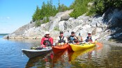 People wearing life vests, hats and sunglasses smiling in kayaks along the water by a rocky cliff with lush greenery