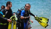 A group of men in scuba gear and wet suits walk through shallow water while holding their flippers