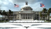 A rectangular building with a raised dome on top and columns with flags in front and cloudy skies above