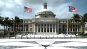 A rectangular building with a raised dome on top and columns with flags in front and cloudy skies above