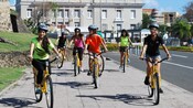 Siete adolescentes en bicicletas recorren una acera con edificios de fondo