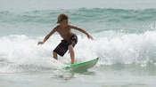 Un niño pequeño con los labios fruncidos se sube a una tabla de surf mientras es empujado por una ola