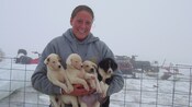 A woman smiles against a snowy backdrop while holding 4 puppies in her arms