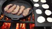 Steaks in a cast iron skillet and pancakes on a griddle cook over an open fire